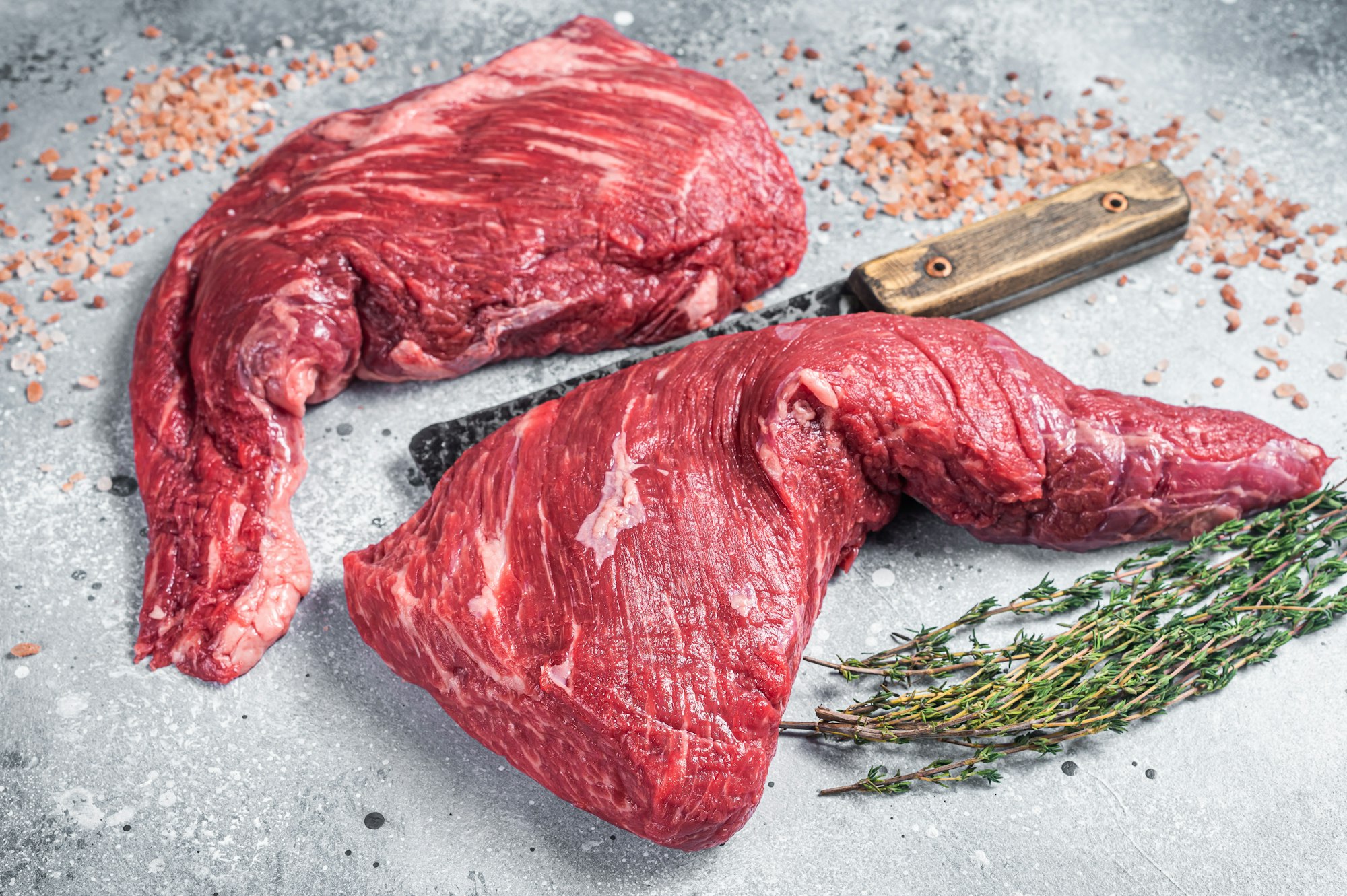 Unccoked tri-tip triangle roast, raw bottom sirloin steak on buther table with meat cleaver.